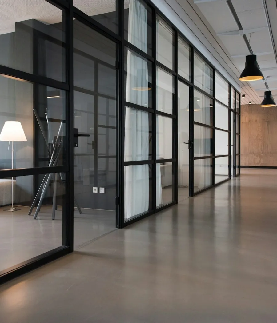 A bright office corridor with glass partitions, wood finishes, and a seating area